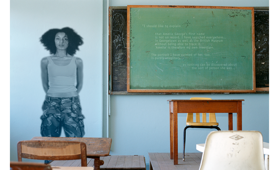 A staged colour photograph of a woman standing in St Roses High School classroom in Georgetown, Guyana with her arms held behind her body. There is a green (blackboard) to her right with text written in white.  She is staring out at the viewer in monocolour. There is a desk and yellow plastic chair arranged on a raised platform in the room with individual desks and chairs in the foreground.