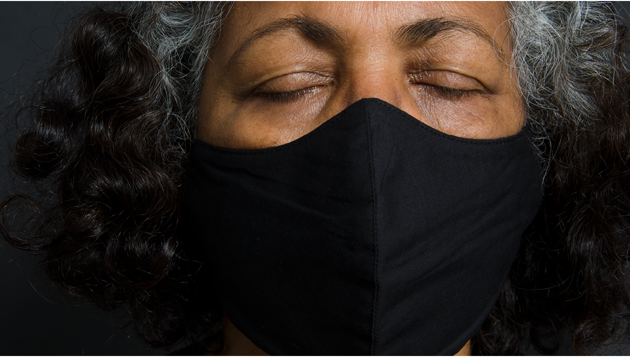  A staged colour photograph taken in the studio using spotlights and a black background. The central plane of the photograph is occupied by a close up self-portrait of the photographer wearing a black mask. Her eyes are closed with the photograph focused on eyebrows, closed eyes, the black mask and some hair. 
