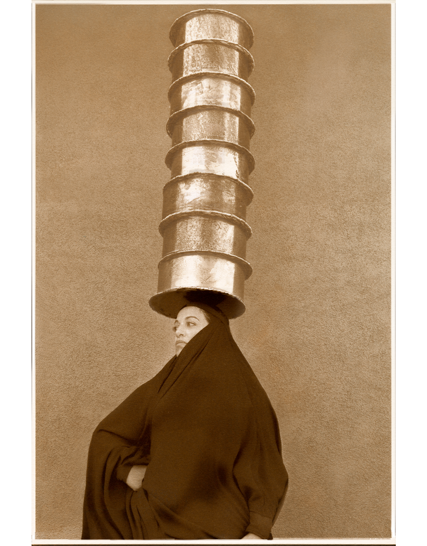 Brown toned photographic portrait of a woman wearing a traditional Iraqi abayya (black garment worn over head and body), with one hand on her hip. On her had, circular pots are stacked over her head.