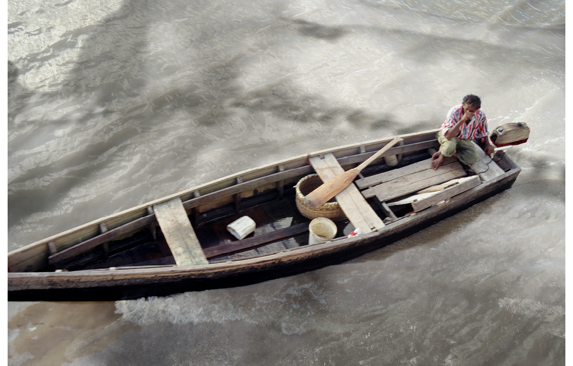 A staged colour photograph of a man driving a wooden boat  using an outboard motor. There is a paddle and plastic containers in the boat. The image is taken from above. He is in the boat of a brown silted river in Guyana. He is wearing a red and white striped shirt. standing in St Roses High School classroom in Georgetown, Guyana with her arms held behind her body. There is a green (blackboard) to her right with text written in white. A black and white shadow appears as a translucent image underneath the boat and stretched across the water. This is an image from an autopsy of a torso of a chest that has been sewn back up post examination. 