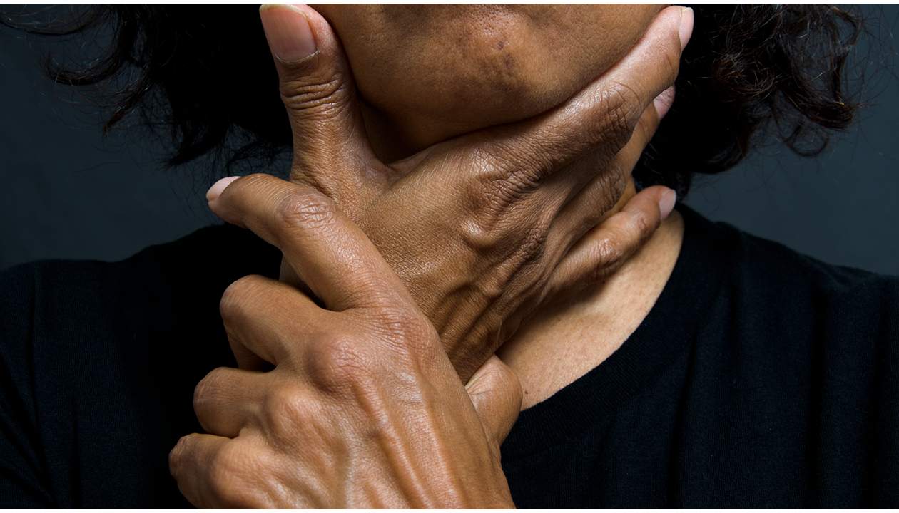 A staged colour photograph taken in the studio using soft spotlights and a black background. The central plane of the photograph is occupied by a close up self-portrait of the photographer's neck and shoulders, her neck being held by both her hands as if caressing it. The photographers chin is visible. The photographer is wearing a round neck t-shirt. 