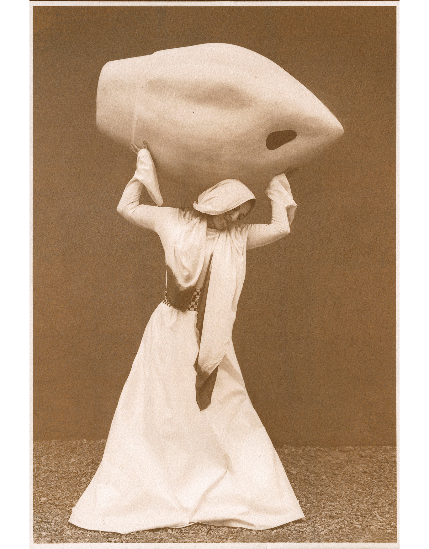 Brown toned photographic portrait of a woman in all white dress, carrying a very large wooden vase with a hole in it over her head.