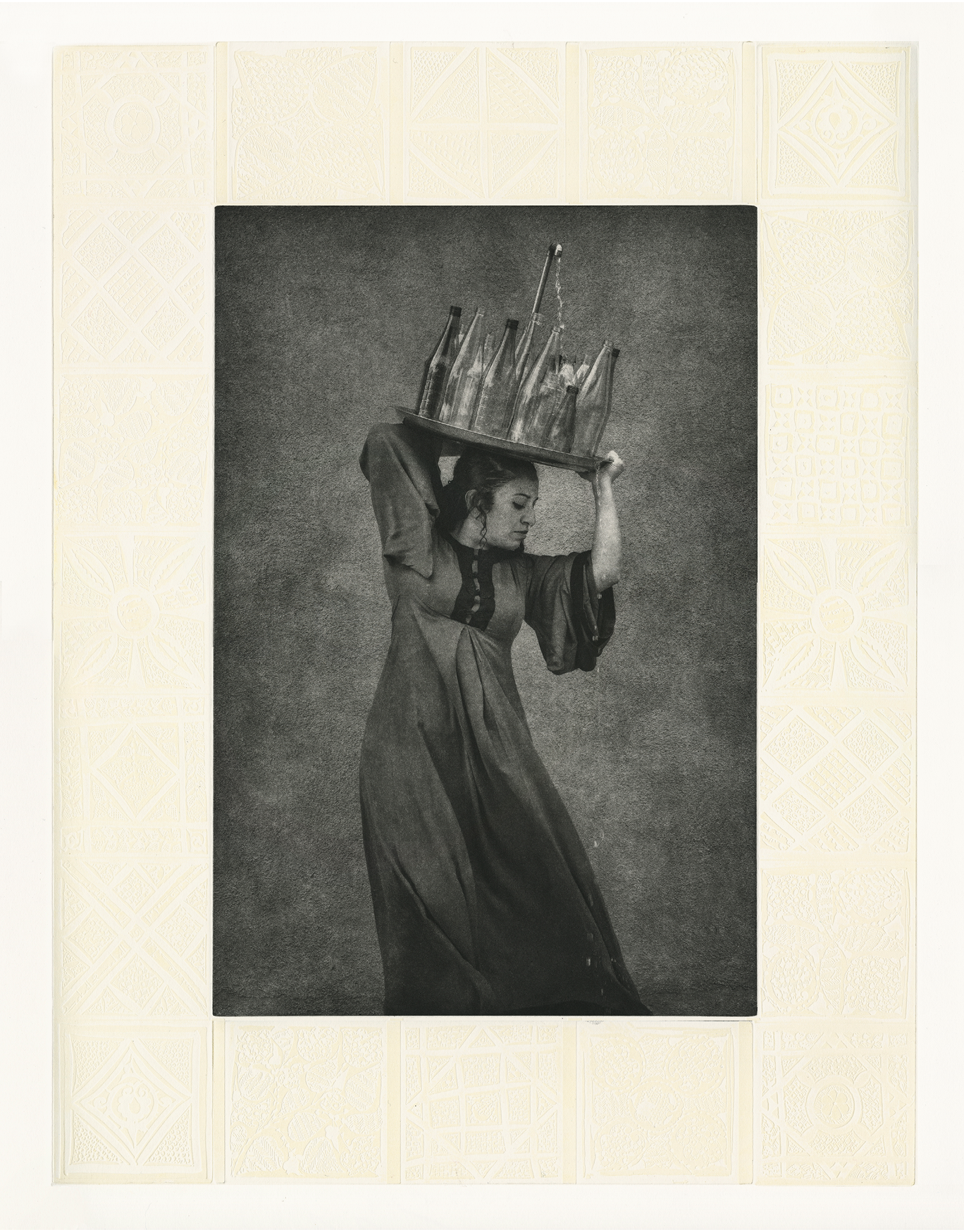 Black and white photographic portrait of a woman in profile view wearing a flowing dress. She is wearing a dress, and carrying a large tray with many glass bottles on top of her head. At the center of the tray is a water spout, with water flowing out and over the bottles towards the ground. The image is framed with an embossed decorative design.