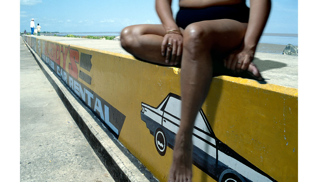 A staged colour photograph of a photograph of a sea wall in Georgetwon Guyana that appears from right to left of the photograph. The sea wall has handpainted illustrations on the front area of the wall. Manipulated onto the seawall is a portrait of someone in black underwear, sitting comfortably with one leg positioned under themselves as if sitting comfortably on the seawall. One bare leg is hanging down and bare arms holding their feet and legs. The upper part of the body and head is obscured from view and cropped away from the photograph. In the far distance visible in the top left of the photograph as the seawall vanishes, are two figures (man and young boy) on the seawall dressed in white shirts and jeans.