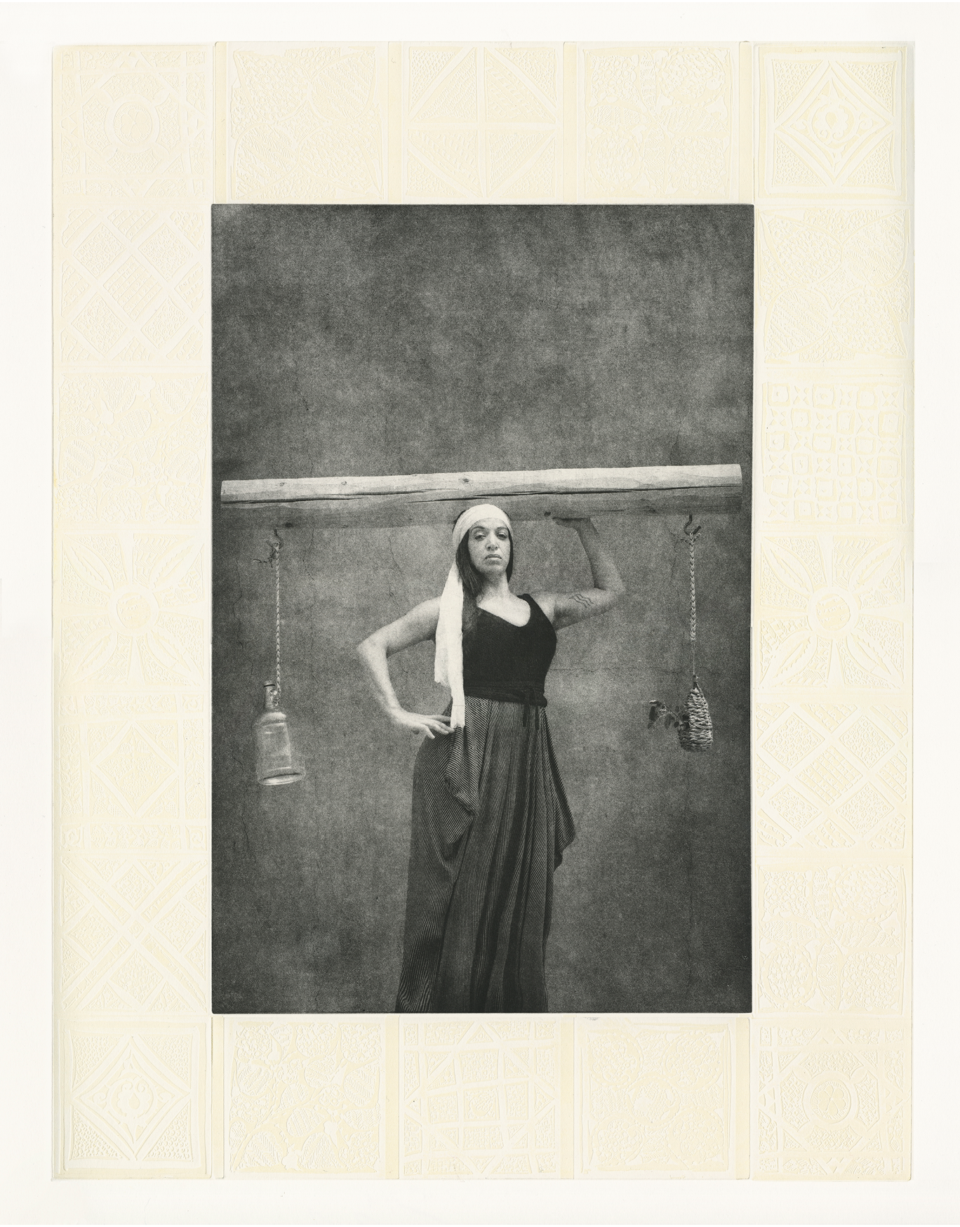 Black and white photographic portrait of a woman, standing with one hand on her hip, and the other hand holding up an electricity pole across her head. The pole appears to be a scale. On one side, a plant hangs from a jute rope, on the other side, an empty glass jug hangs from a rope. The image is framed with an embossed decorative design.