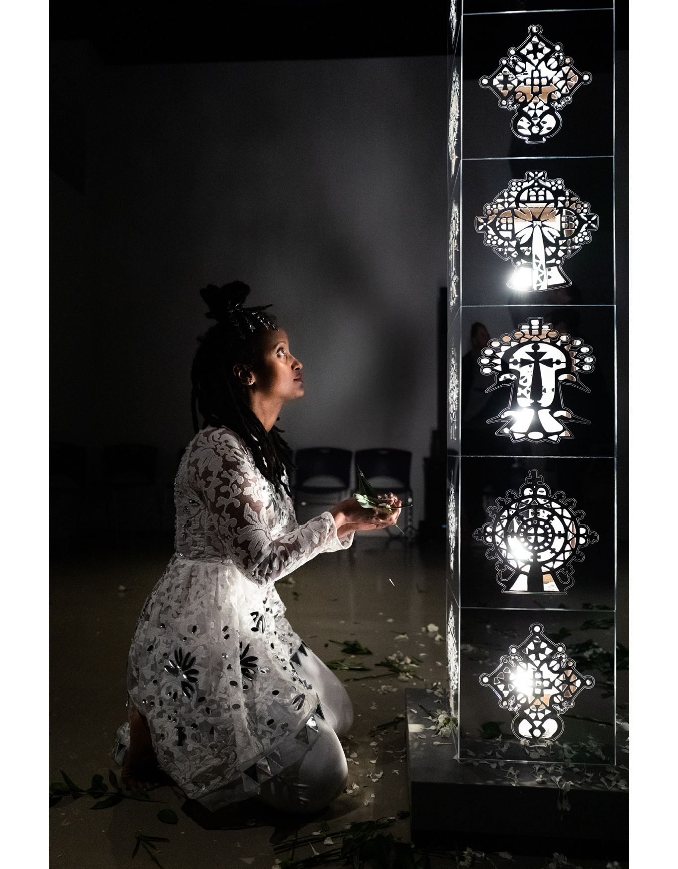 Image of a woman in a white flora lace dress kneels in front of a tall light sculpture with white flowers in her hands. The sculpture is a standing pillar with mirrored square segments. Each square segment of the pillar has abstract cutouts and designs. There is light shining through the cutouts. There are white flower petals on the floor around the sculpture and the woman.