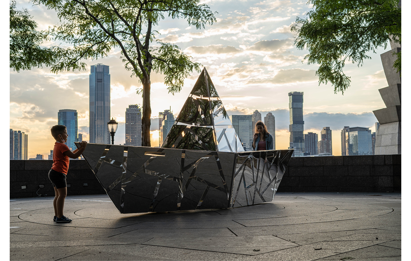 This image shows a young boy holding a geometric mirrored steel boat sculpture. The sculpture looks like a paper boat with lines cut through sections to allow light to pass through remincent of a sundail.