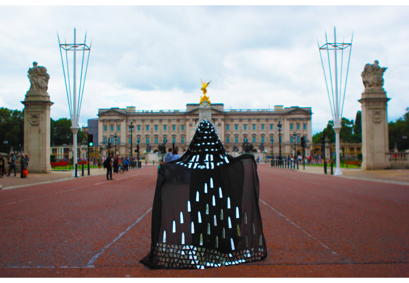 Image of a person standing in the middle the photograph, on a wide road, wearing a long black garment with mirrored patterns. The background of the image shows the entrance to Buckingham Palace, a large buidling with blue windows and two stone statues on either side.