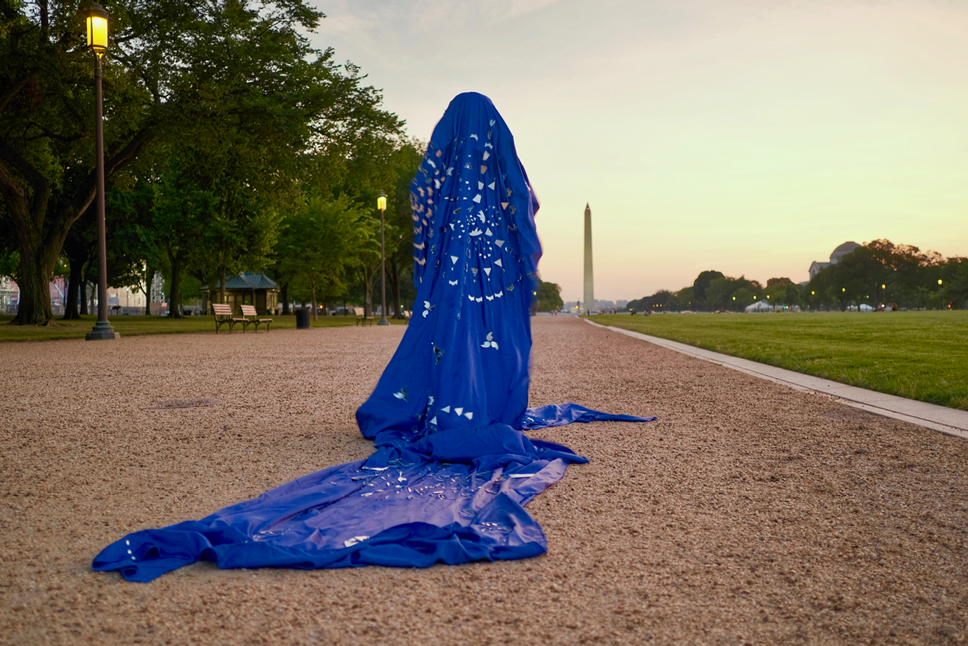 Image of a person standing on a long road. The background of the image shows the Washington Monument, a white obelisk shaped building. The scene is set in the evening. There is a figure standing in the middle of the foreground, in a blue garment with mirrorred patterns and a long train.