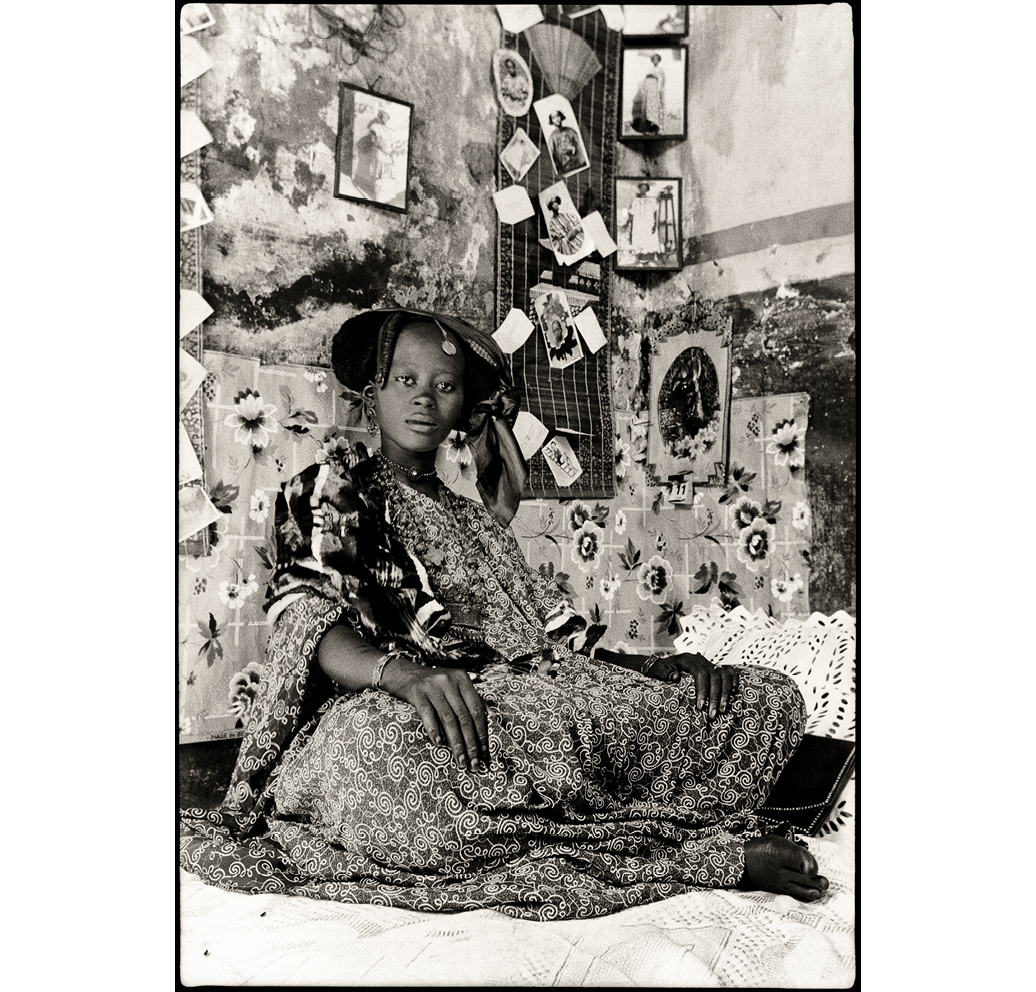 Fig. 1: Black and white image of a Senegalese woman in a long dress robe sitting against a wall that is covered in photographs