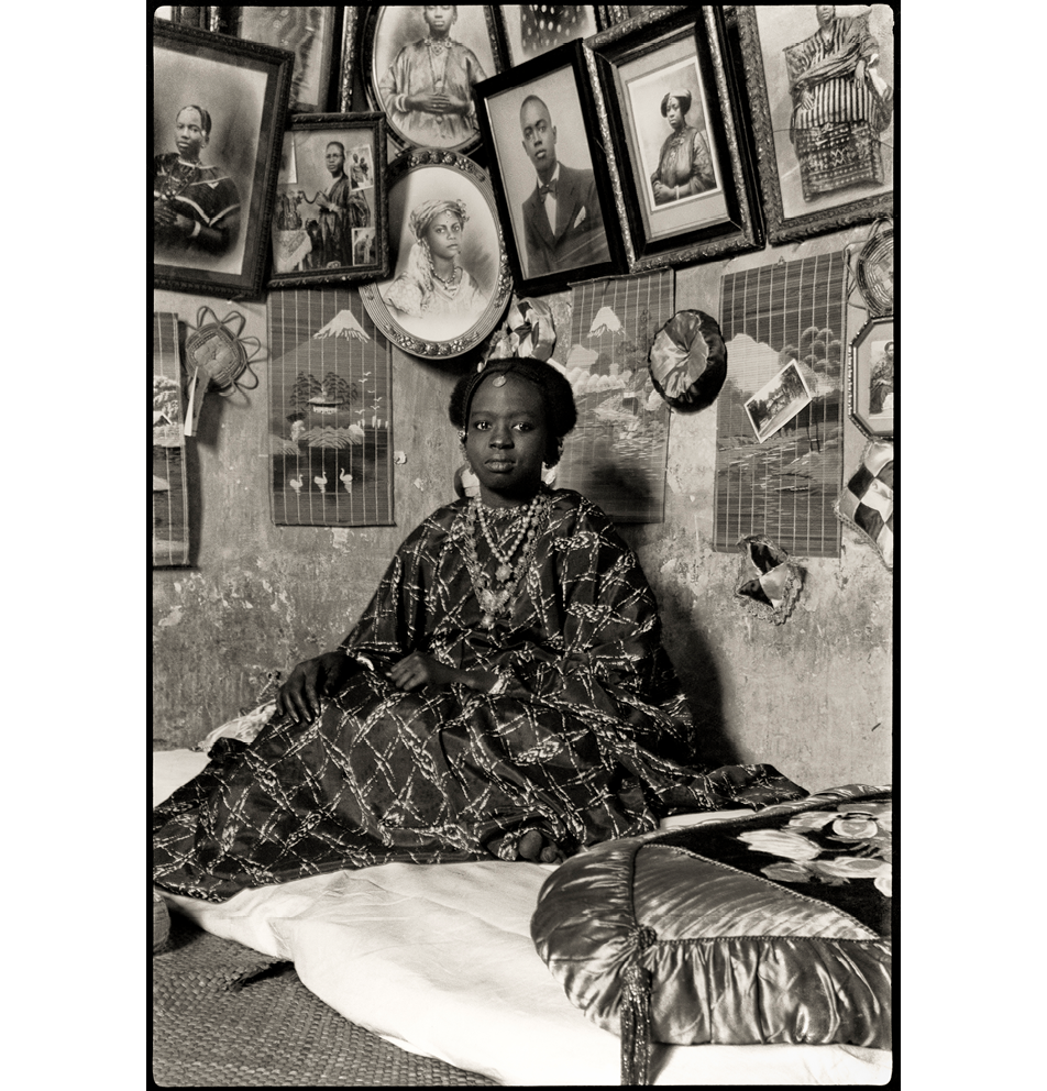 Black and white image of a Senegalese woman in a long dress robe sitting against a wall that is covered in photographs