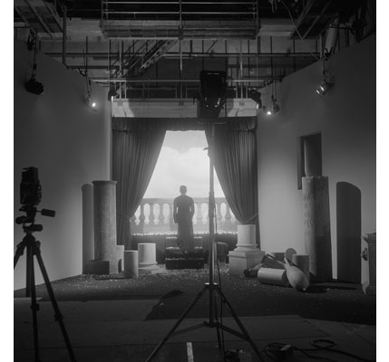 Black and white photograph of a stage set with white walls, a dark floor, and stage lights on the ceiling. A person in a long black dress stands in the middle of the image. They are standing on theater steps in front of a window facing outside column fence. The window has long curtains on either side. The room has prop columns and other objects on the floor. In the image's foreground, there is a camera and tripod on the left side and a light stand in the middle.