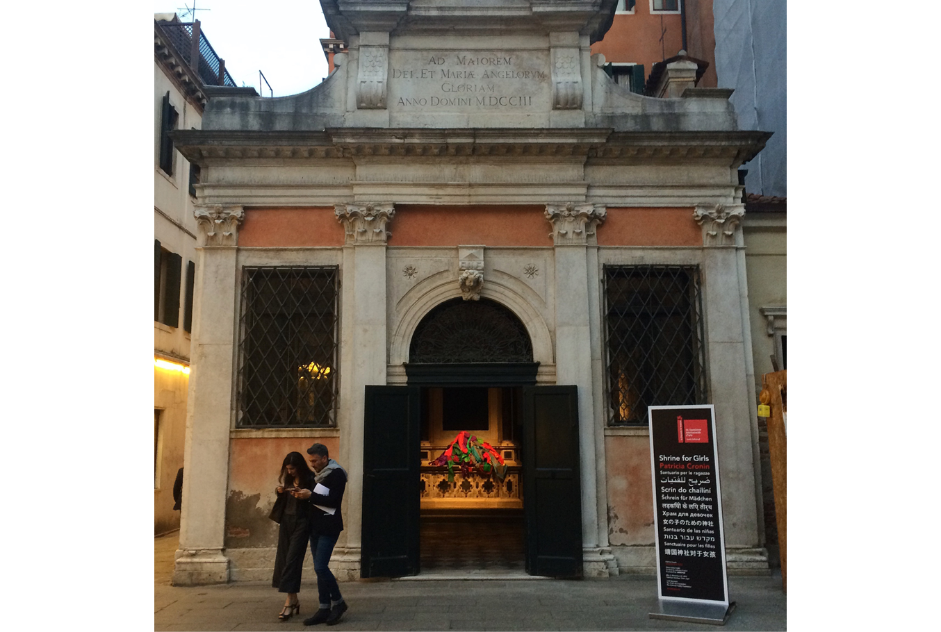 The exterior view of a small orange stucco church with white marble trim in Venice, Italy. The center front green doors are open and the high altar inside is illuminated with some brightly colored objects on top of it. A young man and woman dressed in black are walking away. Signage outside the church for the Venice Biennale reads: 'Shrine For Girls' in English and 12 other languages.