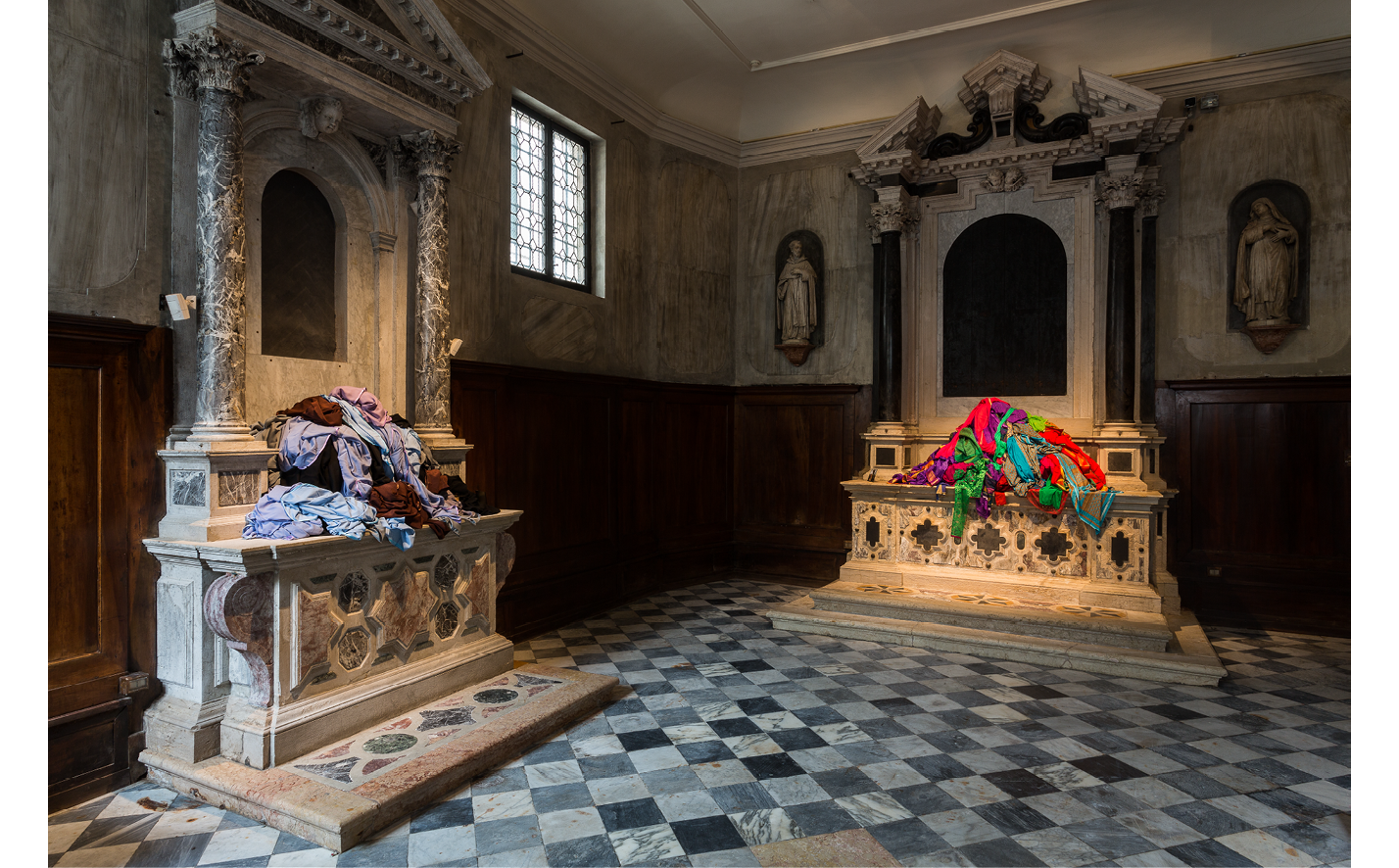 Interior view of the church looking left, with black and white checkerboard marble floor, a stone altar to the left with light blue, lavender, brown and black hijabs arranged on top of the altar. Straight ahead on the stone high altar are brightly colored saris (red, purple, green & gold) arranged carefully.