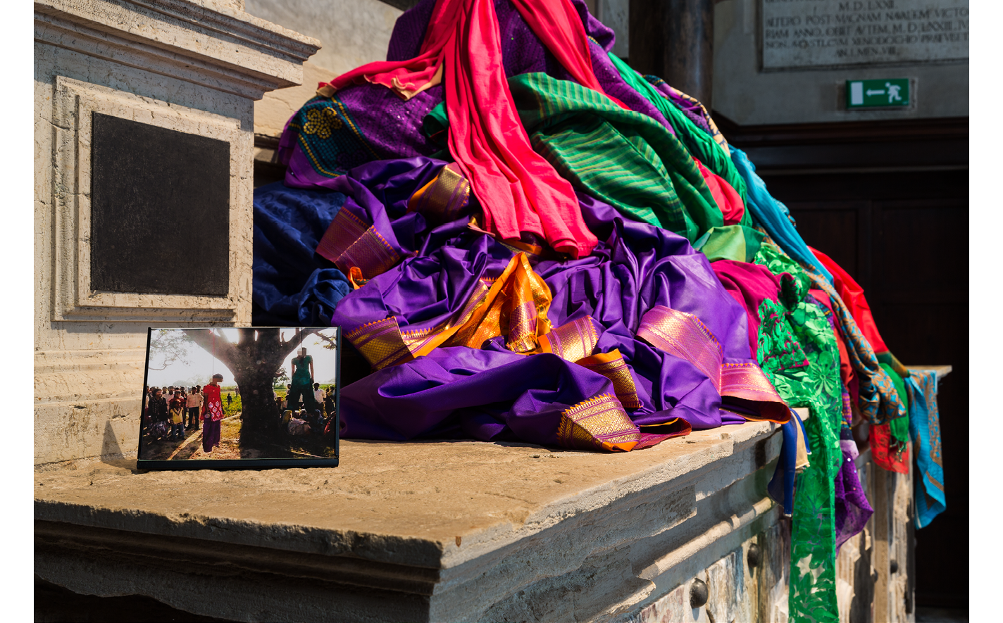 A 3/4 side view of the pile of brightly colored (red, purple, green, & gold) saris arranged carefully on the high stone altar with a small photograph of two lynched girls hanging from a tree facing the viewer.