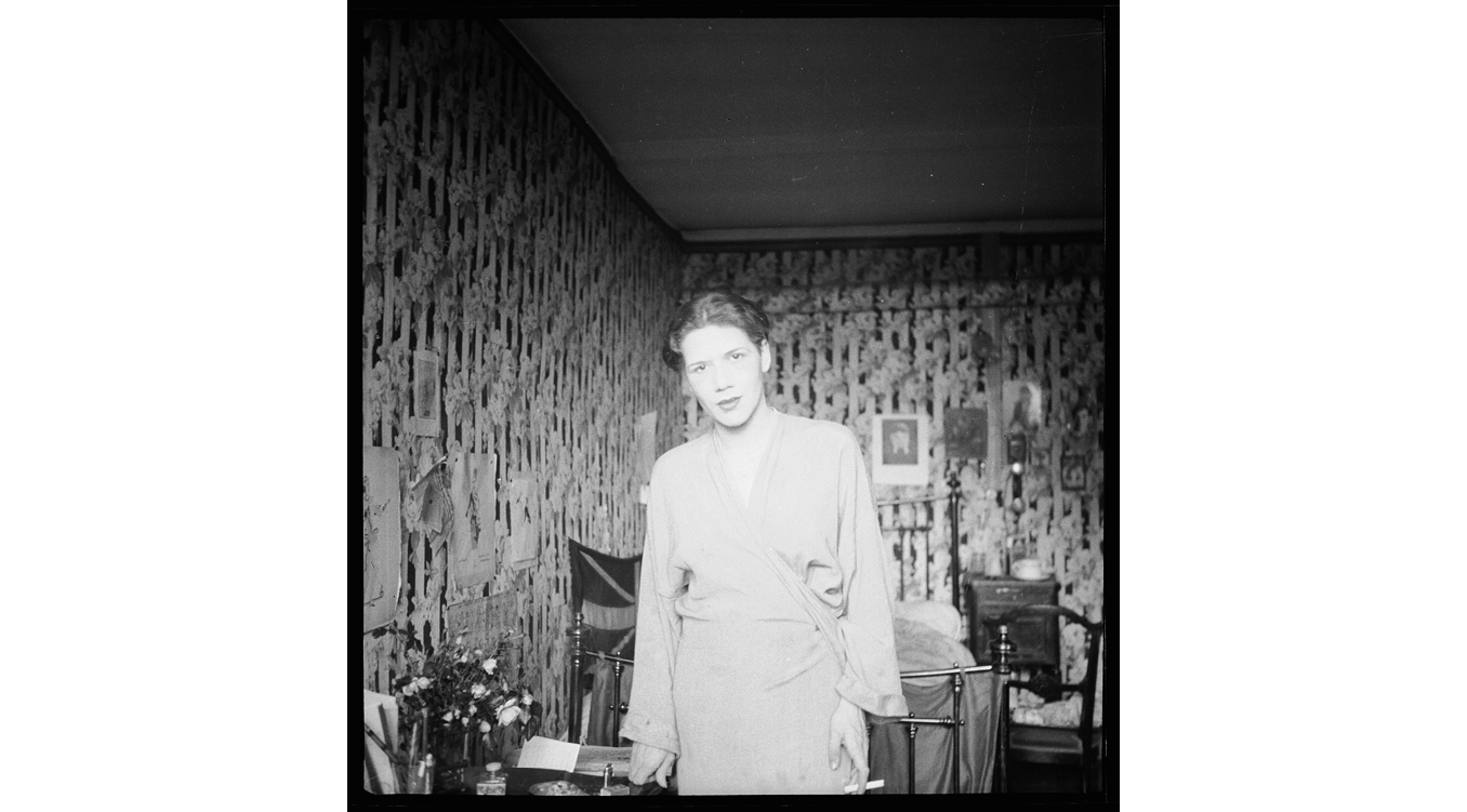 The black-and-white photo from June 1933 depicts Thea 'Mopsa' Sternheim standing in a bedroom filled with furniture and other objects made of wood, brass or steel, and porcelain. Sternheim occupies the center of the image, holding a cigarette and wearing a loose-fitting dress with a V-cut that could also be a robe, her head tilted to one side and her hair pulled back. She has a gentle, inquisitive look on her face. The brightness and evenness of her skin and the similar tone of her garment stand out from the darker, busy surroundings; she inhabits the space almost as an illuminated figure. The walls around her, depicted asymmetrically with two of them receding to a corner just to her left, are covered with striped floral wallpaper, progressively out of focus as it recedes into the background. They are hung with a number of images, including fashion sketches and photographic portraits, that almost disappear into the wallpaper, likewise mostly out of focus. In this space, Sternheim stands amid chairs, a brass bed, and a part of a table or desk covered with papers, writing utensils, and flowers.