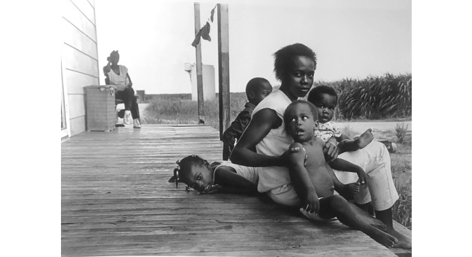 A young woman sits on a porch with four young children behind and on top of her. An older woman sits in a chair behind her in the distance.