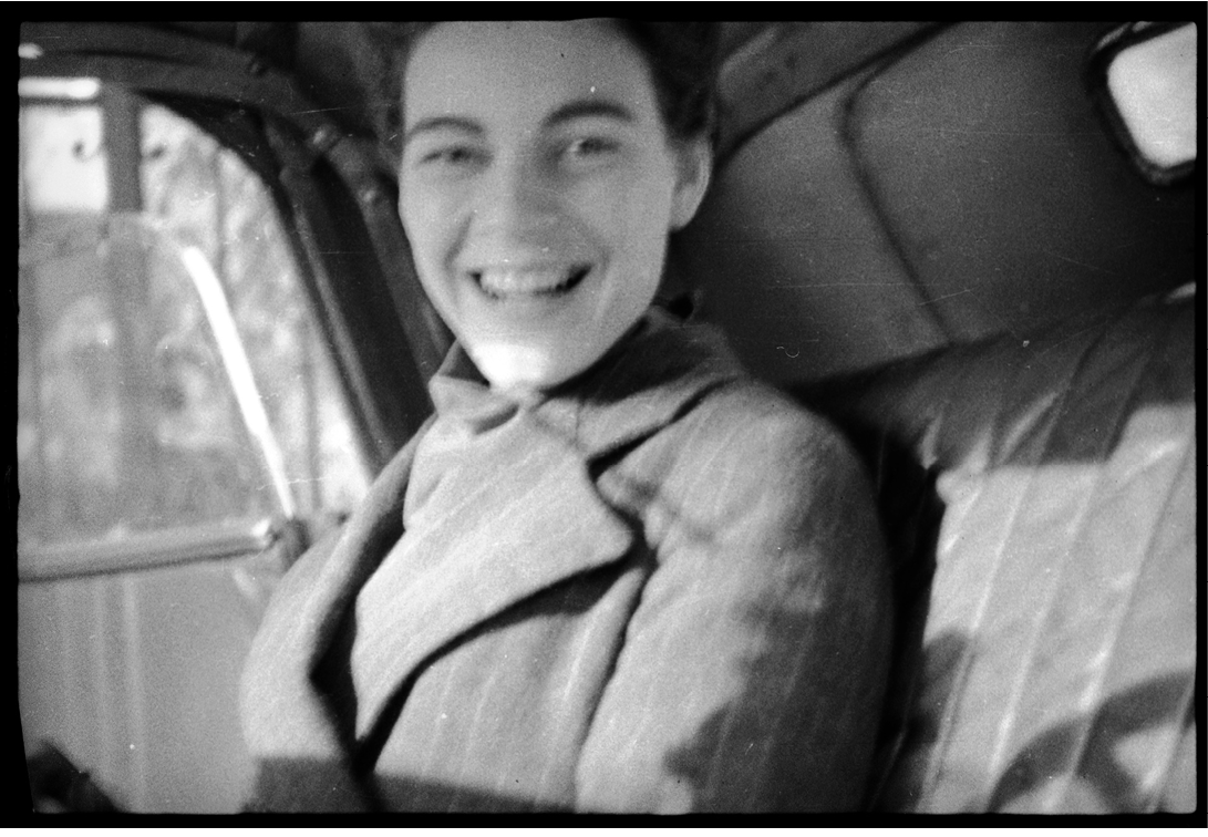 The out-of-focus, black-and-white snapshot from between 1936 and 1938 shows Barbara Hamilton-Wright sitting in the rear seat of a car, an open smile on her face and her eyebrows arching up to mirror the curve of her smile. She is wearing a dark, striped overcoat, with wide lapels, that echoes the stripes visible in the leather or upholstery of the vehicle and in the detailing on the interior panel of the slightly open door. Light streams through the window of the door to her right, illuminating her chest and chin from behind and creating a bright, diagonal stripe horizontally across the photo that casts shadows.