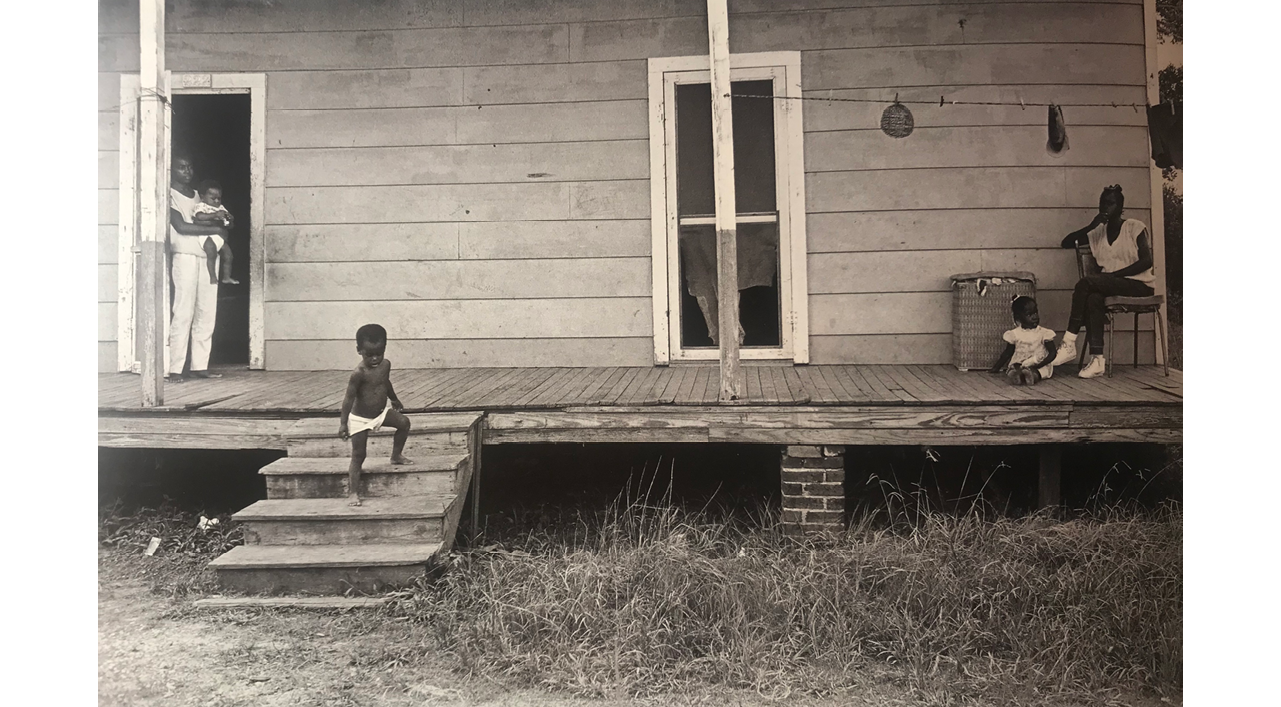 This photograph is a different perspective of the previous scene, capturing the porch straight on. The porch is a straight line across the middle of the composition. A small child walks down  a small set of stairs on the left. The young mother holds one of the children in a doorway behind him. The older woman sits in a chair on the right edge of the picture plane, with a small child at her feet. Both women watch the child on the stairs. 