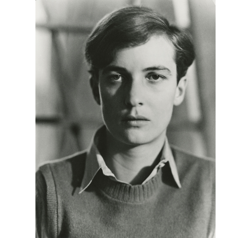 The portrait of Annemarie Schwarzenbach from 1931 is crisp and marked by contrast—one side of her face in light and the other in shadow, the background completely out of focus, producing a diagonally geometric pattern of dark lines, shadowy fields, and bright shapes against the utterly clear features of her face and her clothing. Her eyes seem heavy, intent, brooding, even as they gaze directly ahead, her eyebrows like her lips flat and expressionless, forming a precisely shaped T with the elegant line of her nose. From underneath her finely knit wool sweater, a sharply ironed shirt collar of much lighter, striped fabric opens gently, its curving neckline almost mirroring the sweep of her hair worn short and parted to her left side, in an androgynous style.
