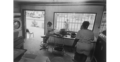 This photograph captures the interior of a laundromat as well as a door and window to the street outside. Inside, a girl folds clothes while her mother watches her. Outside, another mother and child walk by on the street. The scene is book-ended by laundry equipment: washers and dryers. It is sunny and bright outside, and darker inside. 
