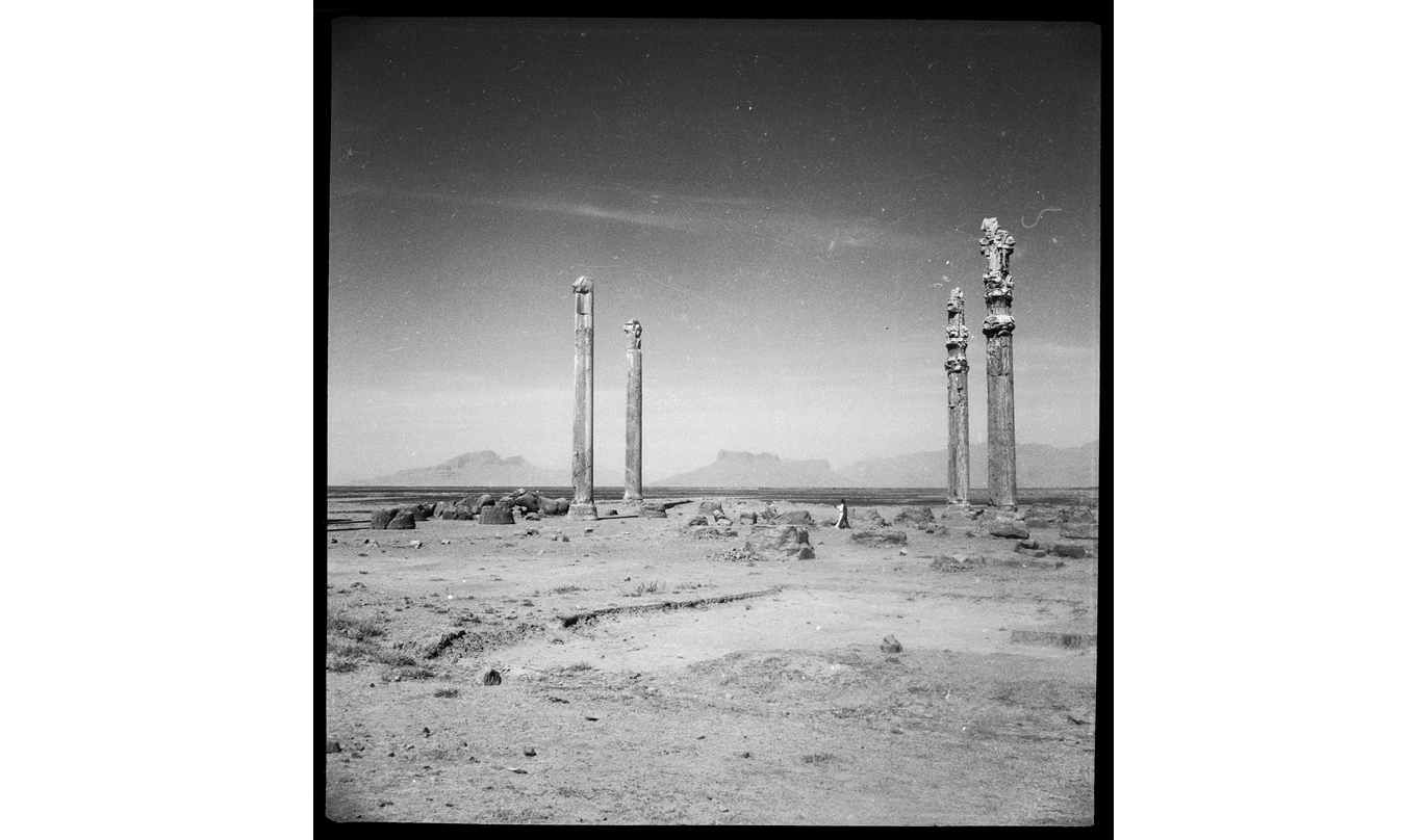 The 1935 photo shows four ancient, ruined columns standing in the desert—once ornate, now in varying states of collapse, as gravity and the weather strip them of their exposed decorative features, leaving only bare pillars of stone. The horizon of the landscape is slightly lower than center, almost classically composed at the bottom third of the image; the sky modulates from a dark well high above, through wisps of clouds and growing light to an almost washed-out silhouette of distant mountain peaks or ridges, jutting up from a nearly perfectly flat plain of uneven rocks and sand. In the foreground, this terrain comes into focus: sparse dry grasses, perhaps bushes, and surrounding the columns, blocks of stone left behind from the temple, and a hollow in the ground that looks like an excavated foundation. A single human figure, likely a man, stands in the center of the columns, so small as to be almost imperceptible, wearing a white robe that is bifurcated into white and black by the light and shadow. Against the sky, the scratches and specks in the photographic print almost appear like stars.