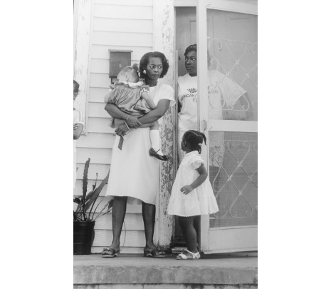 Standing just outside a front door, a mother holds a sleeping child, while another little girl looks up at her. They are all wearing nice dresses and shoes. Inside the screen door an older woman stands in a t-shirt, propping the door open with her arm.