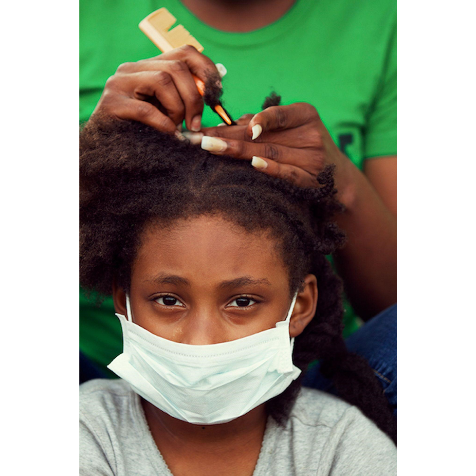 A girl of around ten years of age sits and looks directly at the camera. She has a medical mask on, the type commonly worn during the Coronavirus pandemic. A woman with long, perfectly manicured fingernails stands behind her, doing her hair. The woman's face is not included in the frame. She wears a bright green t-shirt.