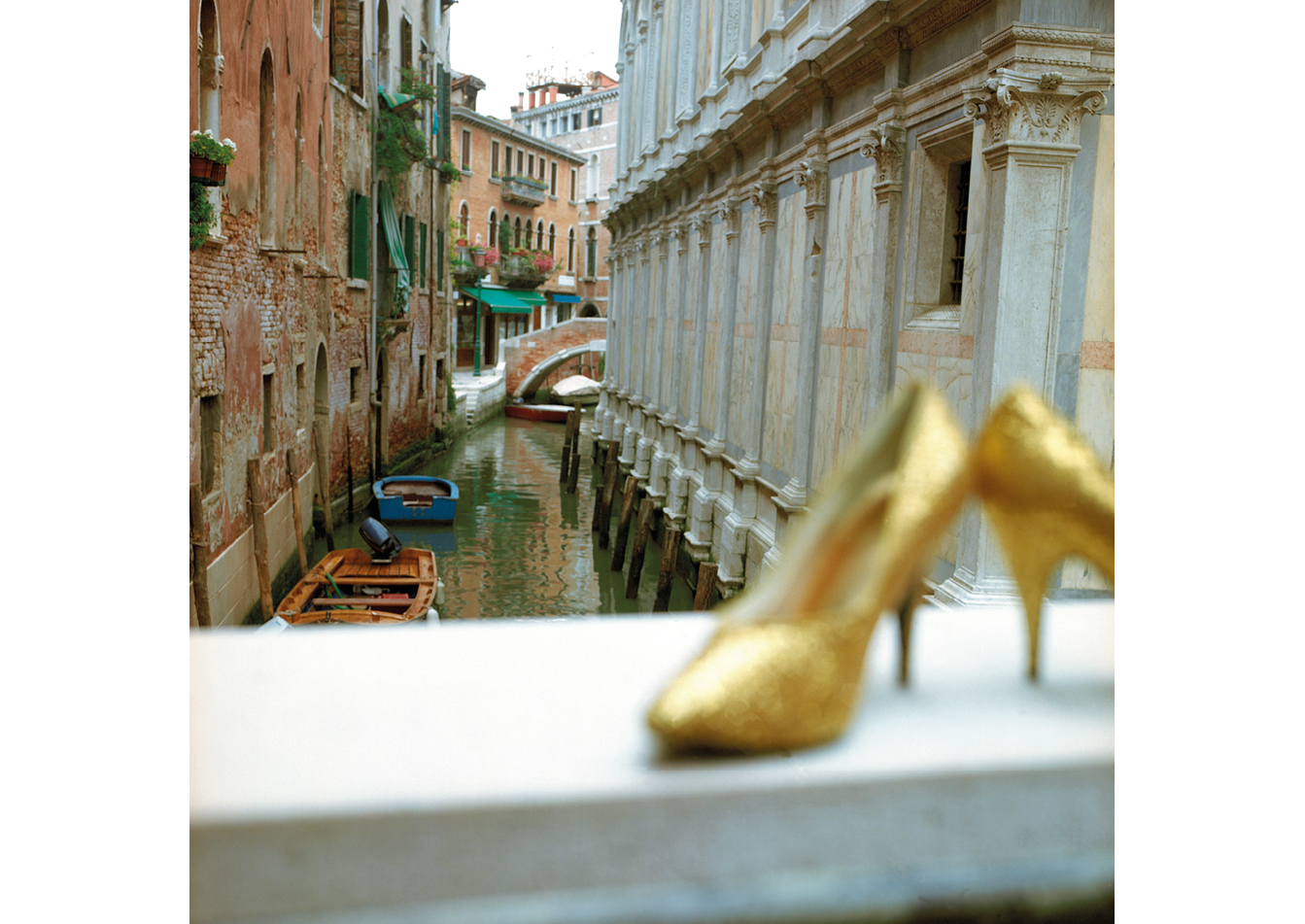 Image of scene in Venice. The background shows two buildings with a canal in in the middle. The foreground shows the ledge of a balcony that has two gold heels rest on top.