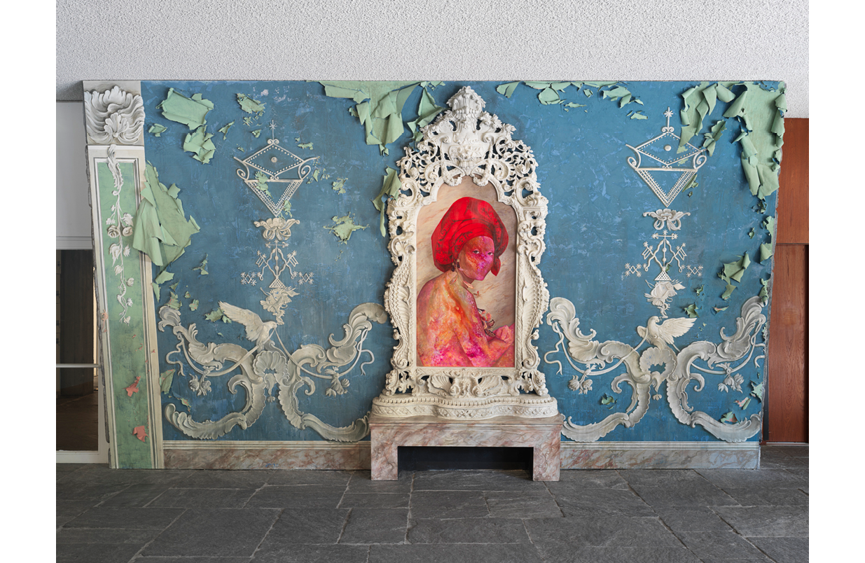 This installation by Firelei Báez includes an ornate decorative frame which sits upon a marble plinth. Inside the frame is a portrait of the Haitian Queen, Marie-Louise Coidavid. She wears a vibrant red tignon and her body is a swirling cosmos of paint. Behind the framed painting is a blue wall with ornate white molding. The wall has fallen into disrepair and the wallpaper has begun to peel, exposing a sea-foam green paper edge.