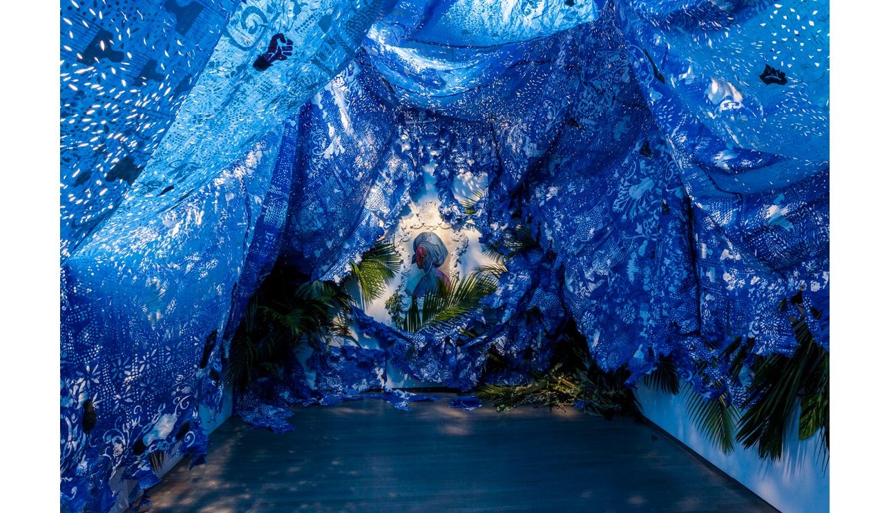 The following image is an immersive installation by Firelei B ez. Blue perforated tarp, which is covered with symbols of healing and resistance and patterning drawn from West African indigo printing traditions (later used in the American South), fills the gallery space. Amidst palm fronts and other foliage an intricately framed portrait of a female figure wearing a cerulean blue tignon looks over her shoulder at the viewer. Her body is marbled with blue, purple, and pink paint and her expressive eyes are the only discernable feature on her face.