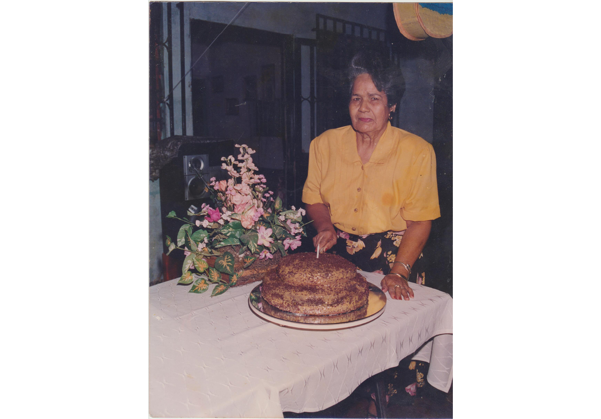 An old woman cuts a cake. She wears a yellow blouse and a black patterned skirt. The table has a white tablecloth and a flower arrangement.