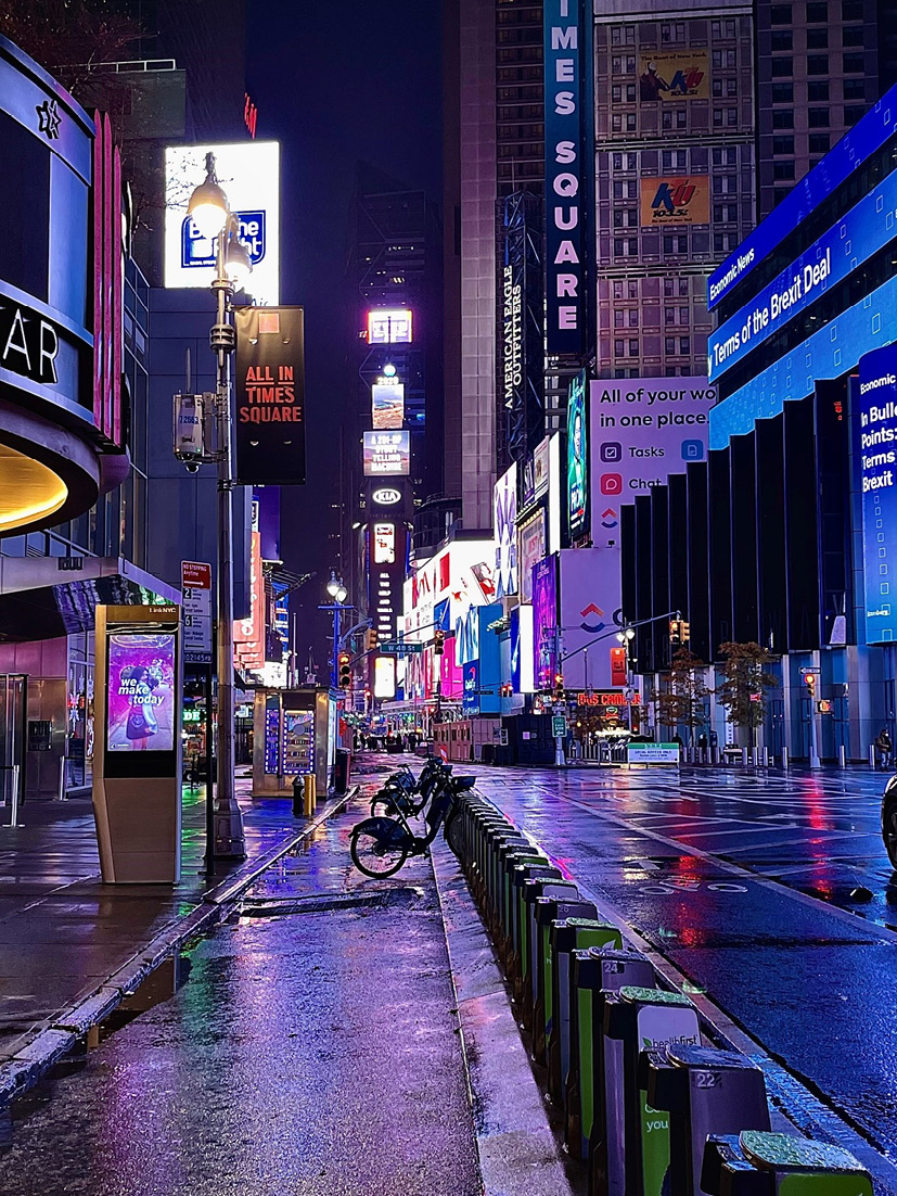 A photograph shows the bright lights of 49th Street in New York at night. There are no people or cars, only a few parked bicyles.