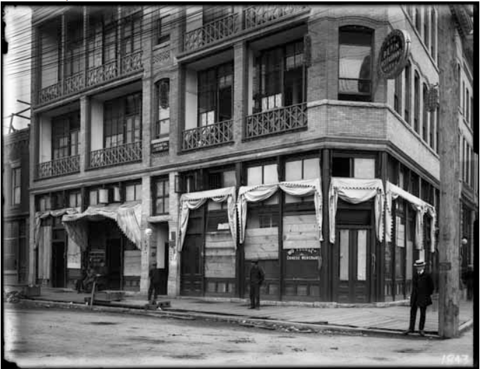 A historical, black-and-white photograph shows boarded-up shop fronts and two bystanders on an otherwise empty street.