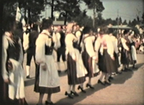 Eighteen folk dancers from Jeppo in traditional costumes dancing their minuet (1977). Partners have just passed each other standing back to back, screenshot by Gunnel Biskop after Kim Hahnsson’s film.