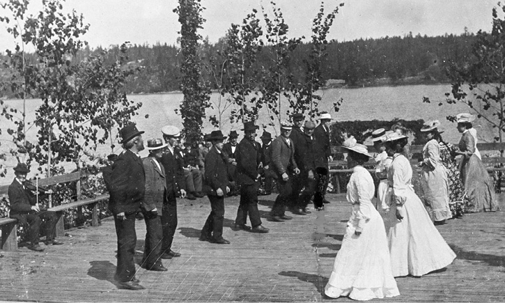 Folk dancers from Kimito dance their minuet at a summer party (1904). The transition steps are taken, and the ladies and gentlemen have turned on opposite sides, at the front end of the row among the men dances school teacher Nils Oskar Jansson, photograph by Otto Andersson.