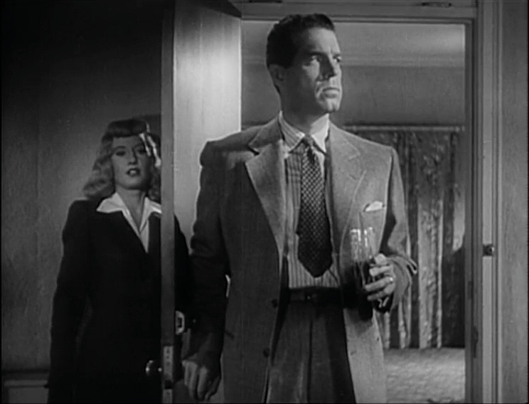 A screenshot taken from a black-and-white film. A man in a light suit stands at an open door with a half-full glass in his left hand and his gaze to his left. A woman with light-coloured wavy hair, a black blazer and a white shirt stands behind the door. Beyond the open door, a room and thick curtains are visible.