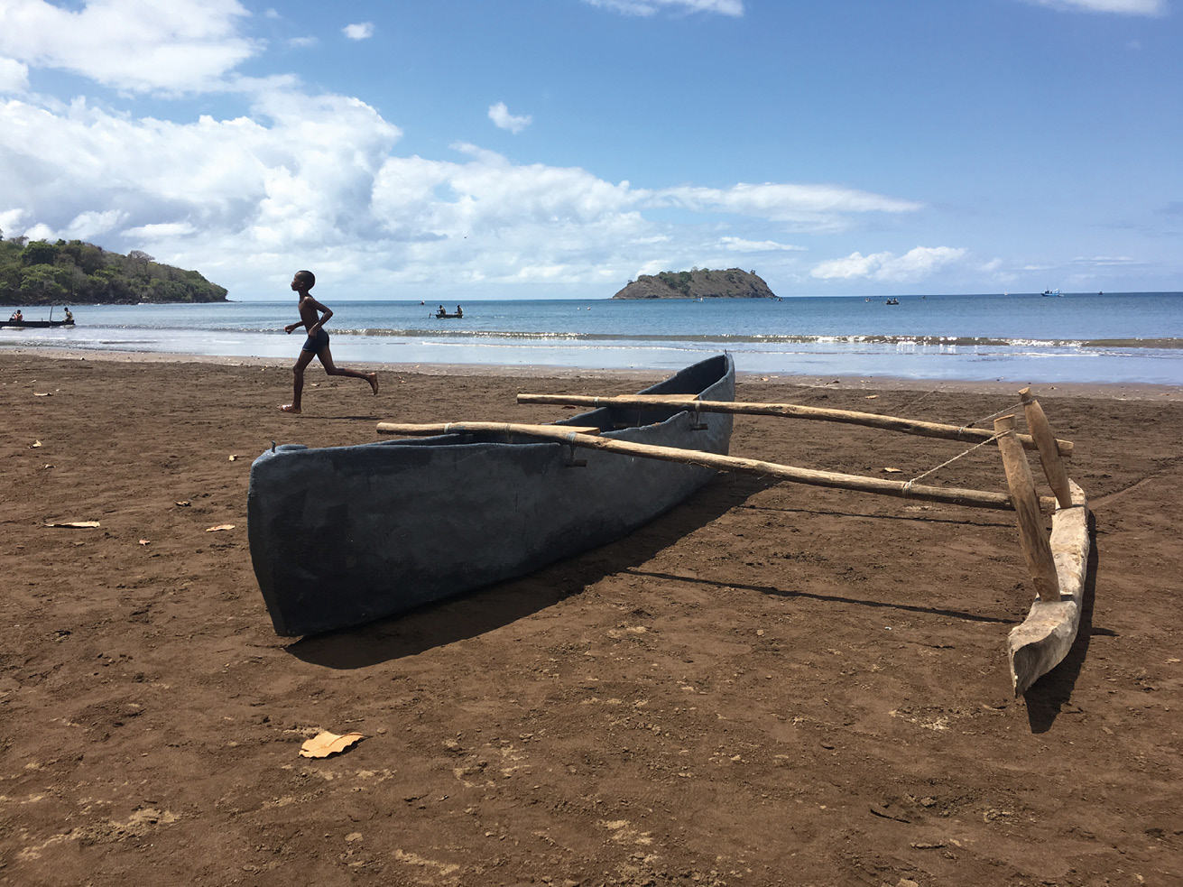 A young boy with a swimsuit runs on an earthy shoreline where a blue Pirog boat lies, a banana-shaped boat with a single wooden outrigger on its right side. In the background is the light-blue ocean with two small green and brown islands.