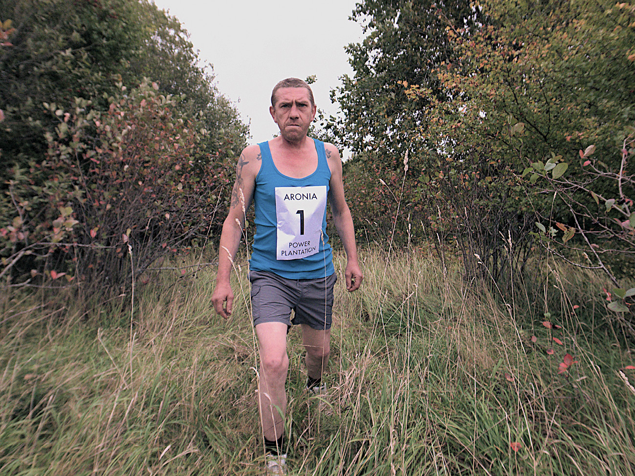 A man is standing in between plants of a re-wilding plantation in the west of latvia. He is asked to imitate whilst standing still the movement of a marathon runner. He is dressed fakingly as a runner, with second-hand clothes: dark grey shorts; blue sleeveless shirt, black socks and running shoes. He has a paper print with number one on his chest. He looks directly in the photocamera with a severe expression, emphasised with his lavishley tattood arms and very short cut hair. It is the first of 42 photos, as in 42 kms of a marathon. The idea of the photo series originates from the idea that a marathon at the plantation would have kept the overtaking grasses down.",