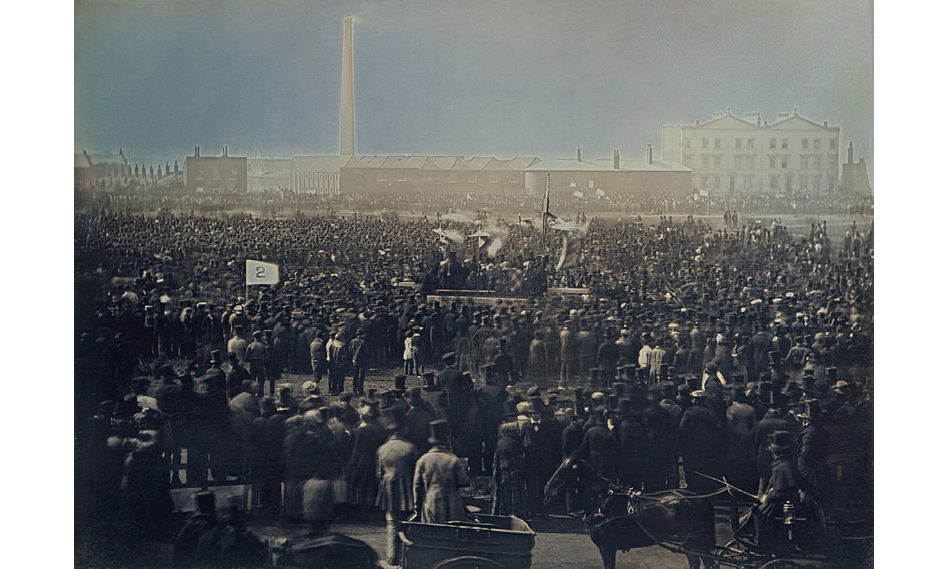 A daguerreotype photograph from 1848 London showing the Great Chartist Meeting at Kennington Common: a crowd of supporters gather around a platform with speakers; in the background, a tall factory chimney and a large house to the right