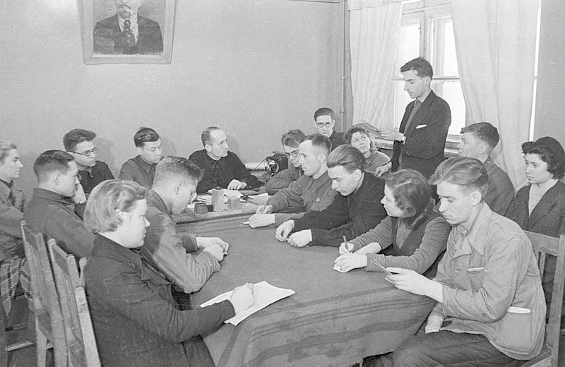 A black and white photograph from 1932 of a Komsomol meeting at the Magnitka plant: you men and women sit down around a table with pen and paper in hand while one young man stands and speaks; in the background a wall with a portrait of Lenin and a closed window with white curtains.