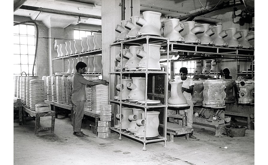 A black and white photograph ca. 1960-1970 of a bathroom fixture factory in the Netherlands: shows three workers preparing bathroom fixtures to be baked in tunnel ovens; throughout the room, individual parts are stacked in shelves.