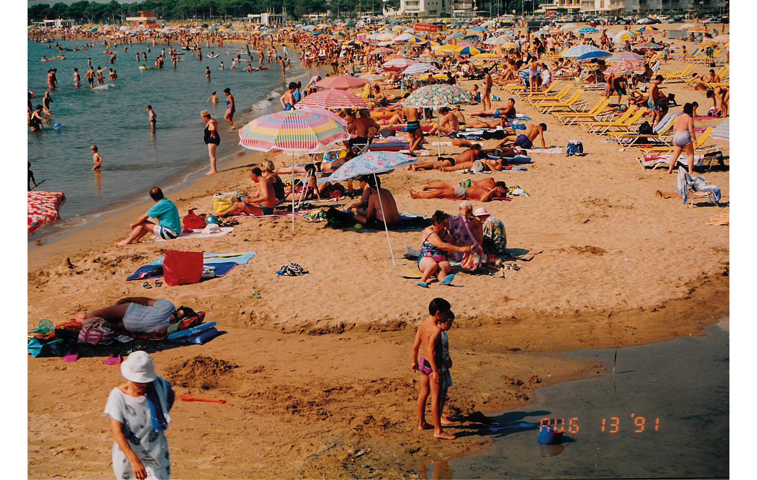An in-color photograph from 1991 Spain of tourists in Costa Brava: shows a crowd of tourists both in the water and gathered around the coast; in the sand, an array of colorful beach towels, umbrellas, and chairs