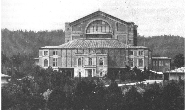 A black and white photograph from 1882 of the Bayreuth Festival Theatre shows the exterior and front-facing side of the theater and its surrounding forests.
