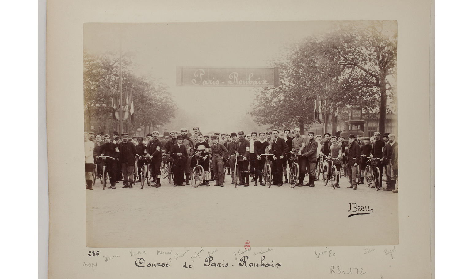 A black and white photograph from 1896: shows participants of the Paris-Roubaix cycle lining up at the starting line; in the background, trees on each side of the road and a banner written â€œParis-Roubaixâ€�; at the bottom, a handwritten inscription reading â€œCourse de Paris-Roubaixâ€�.
