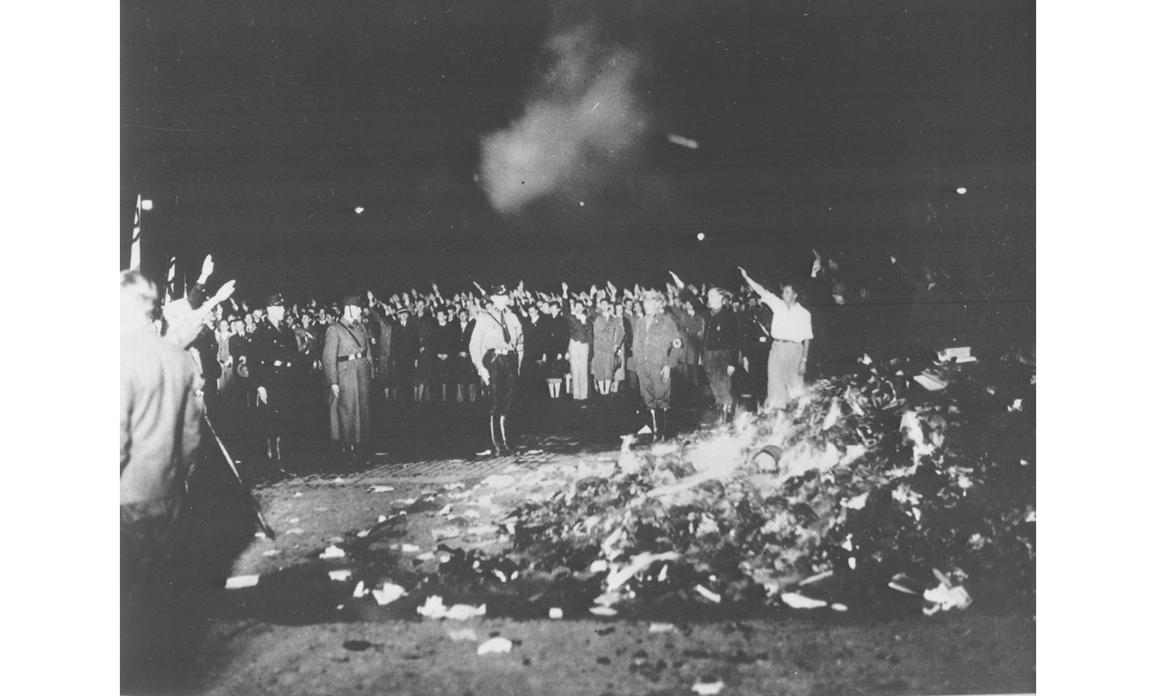 A black and white photograph from 1933 Berlin: captures a book burning by students on Berlin’s Opernplatz; a large crowd of students circle a pile of burning books while holding the Nazi salute; in the background, white smoke emanating from the pile rises against the night sky.