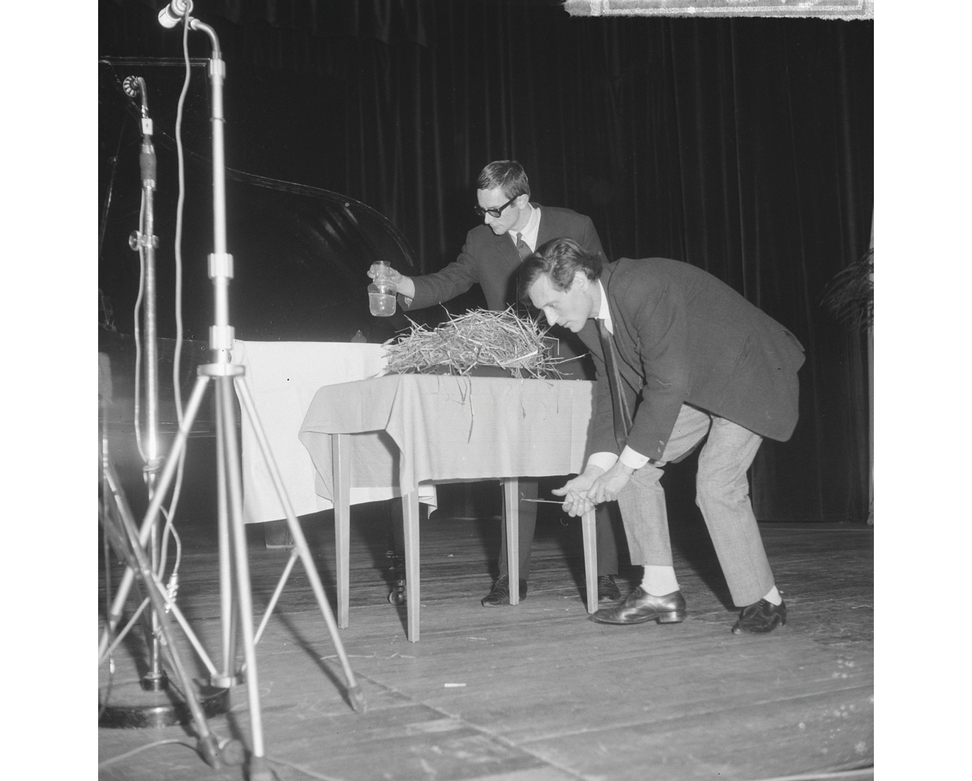 A black and white photograph from 1964 depicting a Fluxus concert at Kurhaus Scheveningen: two Fluxus artists attempt an experiment with a table full of straw and an unknown liquid; to the left rest two microphone stands.