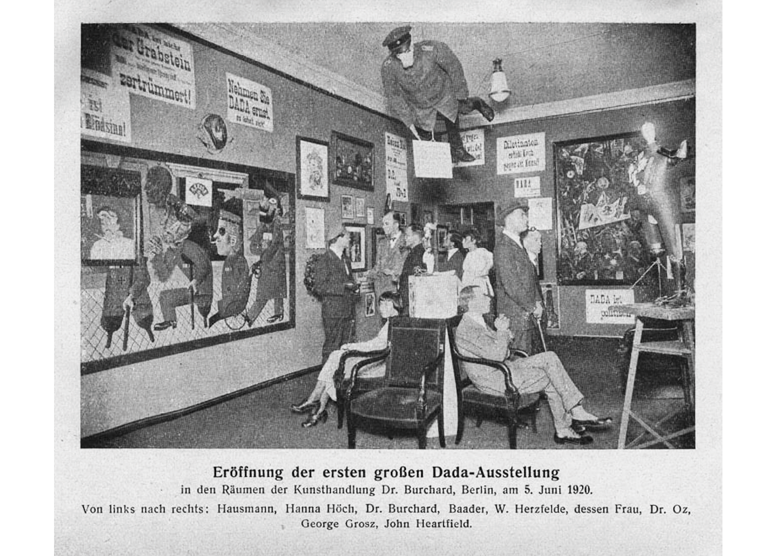 A black and white picture of the grand opening of the first Dada exhibition in 1920 Berlin: in the center, an effigy of a German officer with a pig’s head hangs from the ceiling over a circle of chairs; exhibition visitors enter the room, and observe the various exhibition pieces; on the walls are various art pieces such as painting, drawings, pictures, and posters written in German; on the bottom a title and description of the photographs and names of visitors written in German.