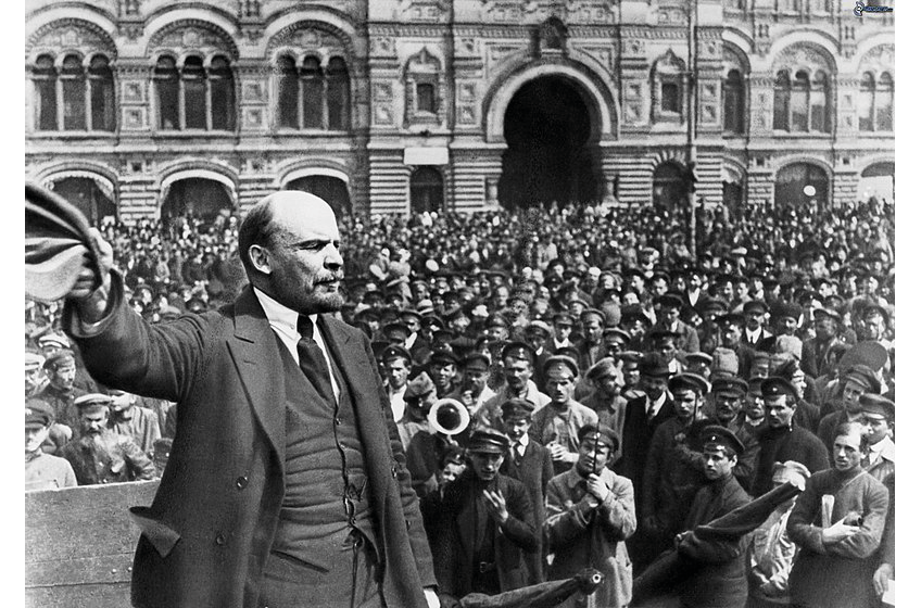 A black and white photograph of Soviet leader Vladimir Lenin giving a speech: shows Lenin waving his hat as he stands in the middle of a crowd of servicemen in the middle of the Red Square.