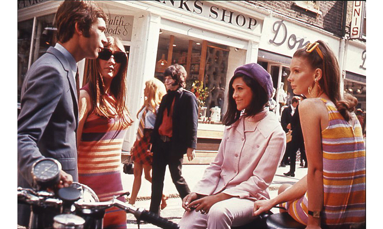 An in-color photograph from 1966 of young adults in London’s Carnaby Street: three young women dressed in colorful clothes and one young man chat in the middle of a busy street; in the back, a couple walks through the street; in the background, names and window panes of various retail stores.